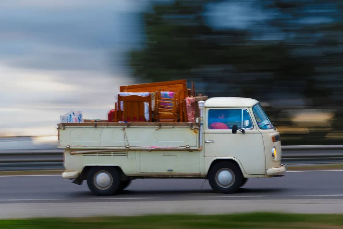 An old pickup loaded with luggage speeds along a highway, blurred by motion.