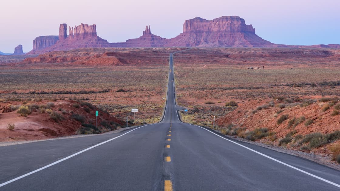 A straight two-lane highway running toward the red buttes of Monument Valley under a pink dawn sky