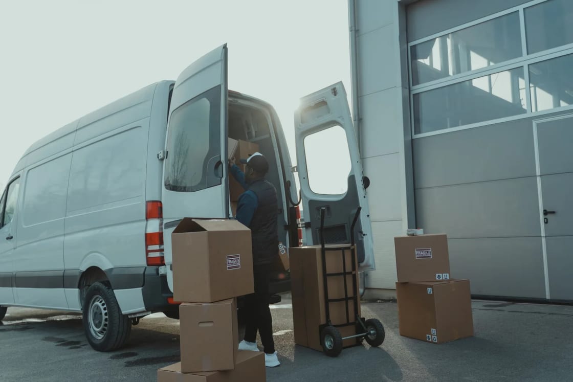 A white van with its back doors open beside stacked moving boxes and a hand truck.