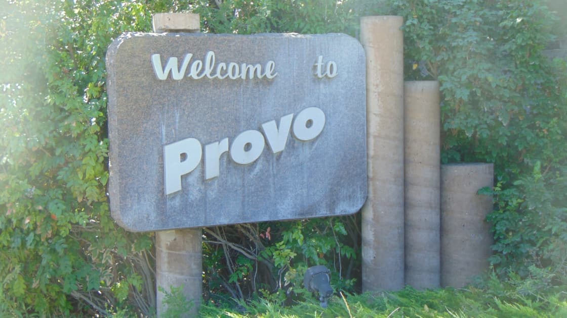 A weathered Welcome to Provo sign in raised letters beside stone pillars, framed by green shrubs