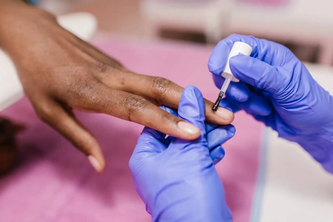 A technician in purple gloves applying polish to a client's fingernail at a pink salon table