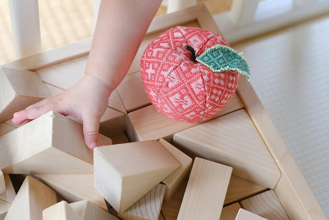 A toddler's hand reaches into a tray of plain wooden blocks beside a fabric apple, the kind of play a sitter sets up at home.