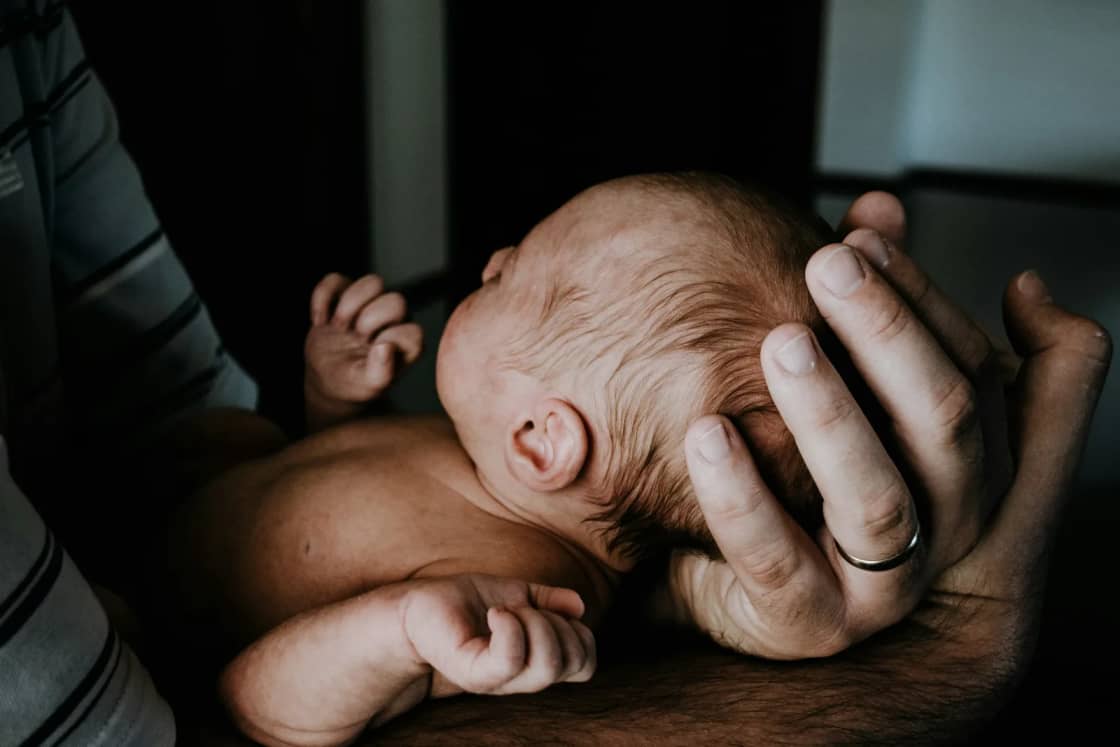 A father cradles a newborn against his chest, one hand supporting the baby's head, his wedding ring visible in low light.