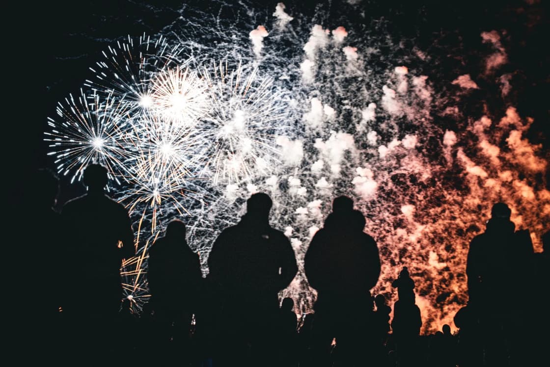 Silhouettes of a crowd watching gold and white fireworks burst overhead against a dark night sky