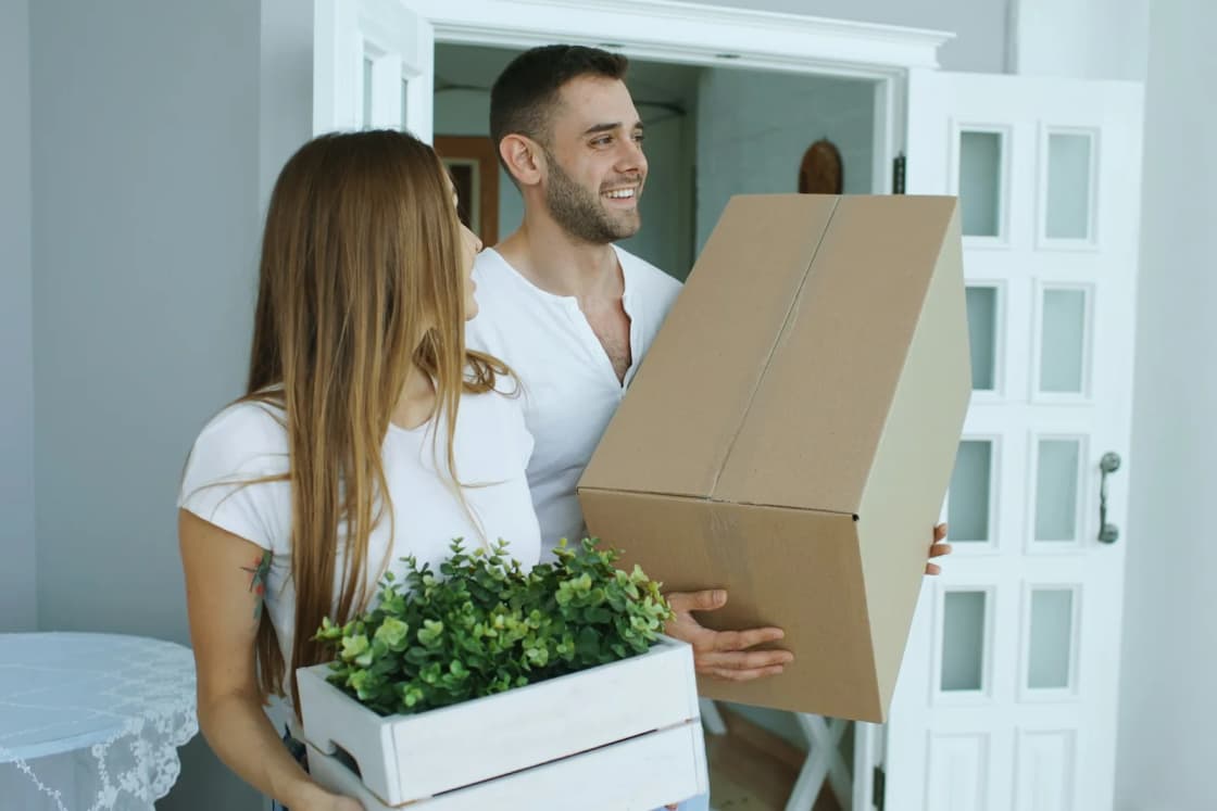 A smiling young couple carries a cardboard moving box and a crate of potted greenery through the door of their new apartment.