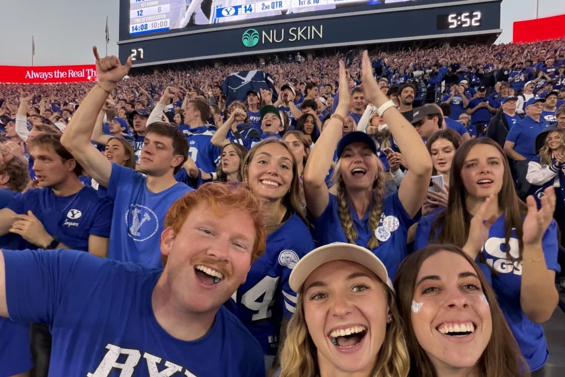Smiling BYU fans in blue take a selfie in the student section, with the stadium scoreboard behind them showing BYU 14 in the second quarter
