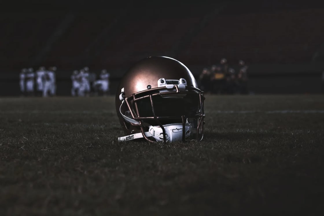 A football helmet resting on the grass of a dark field at night, with players blurred in the distance