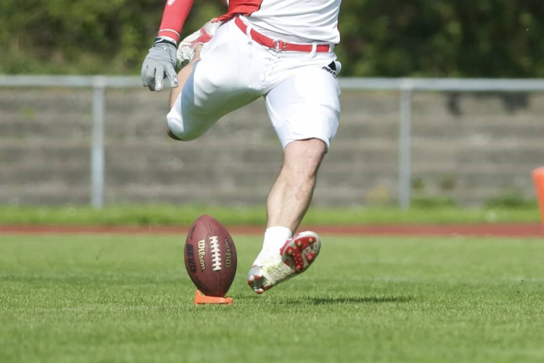 A kicker's leg swinging toward an American football standing on an orange kicking tee on grass