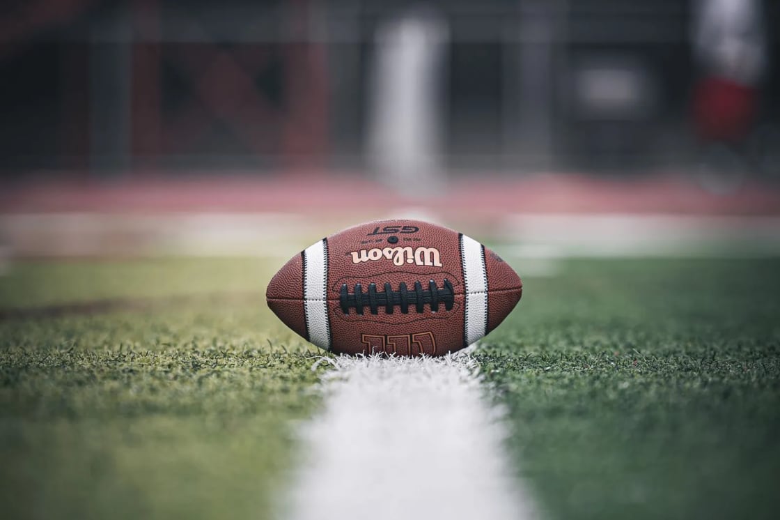 A football resting on a white yard line of a turf field, with empty stands blurred behind