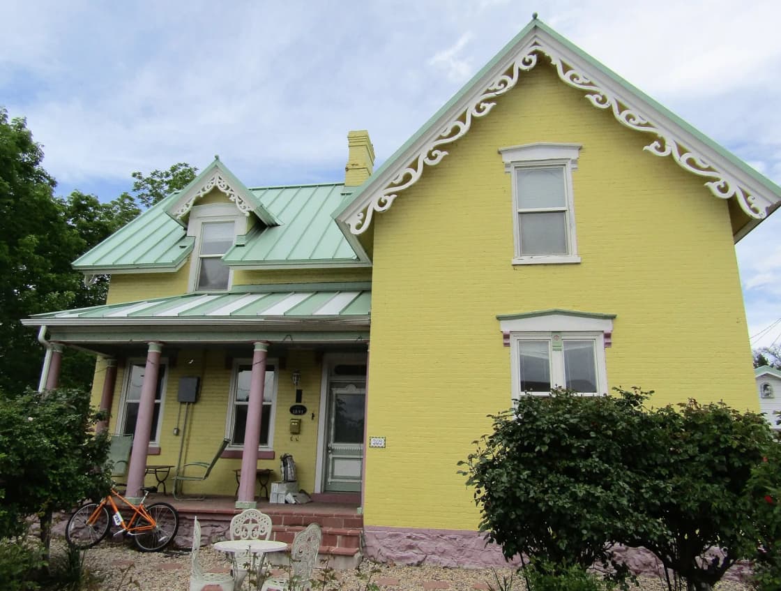 A yellow two-story brick house with white gingerbread trim and a green metal roof on a tree-lined street in Provo, Utah