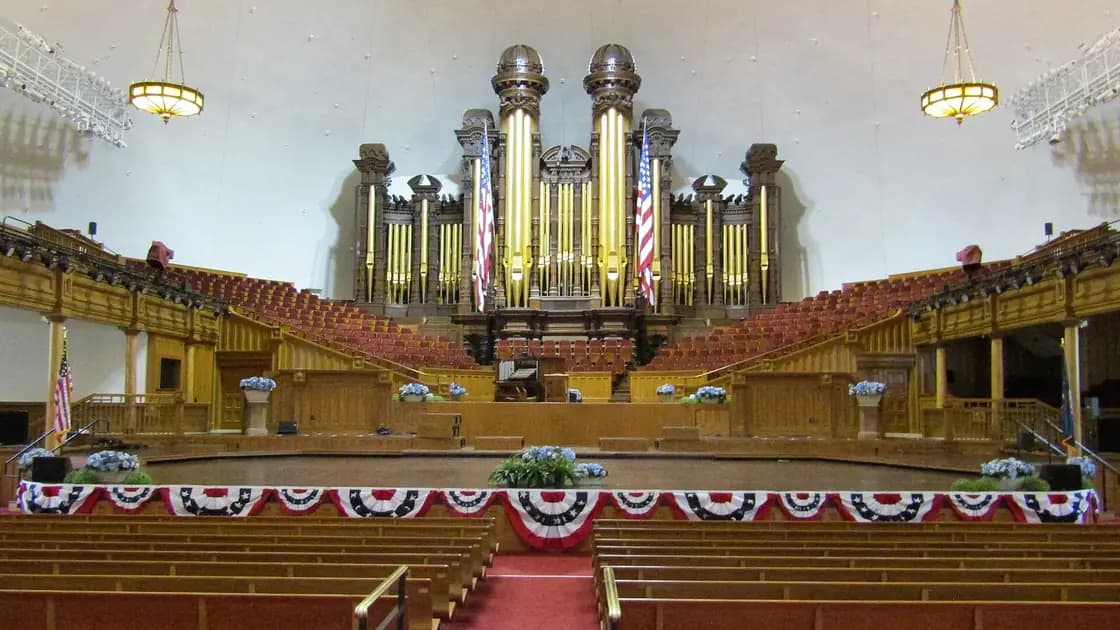 Empty pews facing the gold pipe organ inside the Salt Lake Tabernacle, with American flags and red, white and blue bunting at the stand