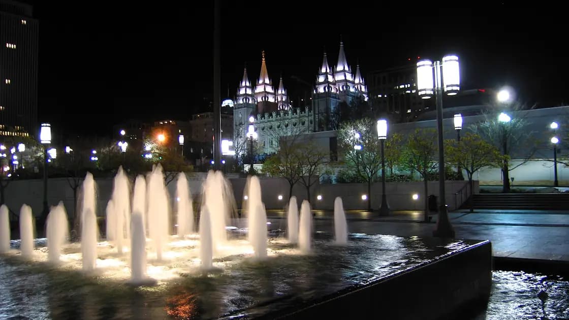 Fountains lit up at night in the foreground, with the floodlit spires of the Salt Lake Temple behind