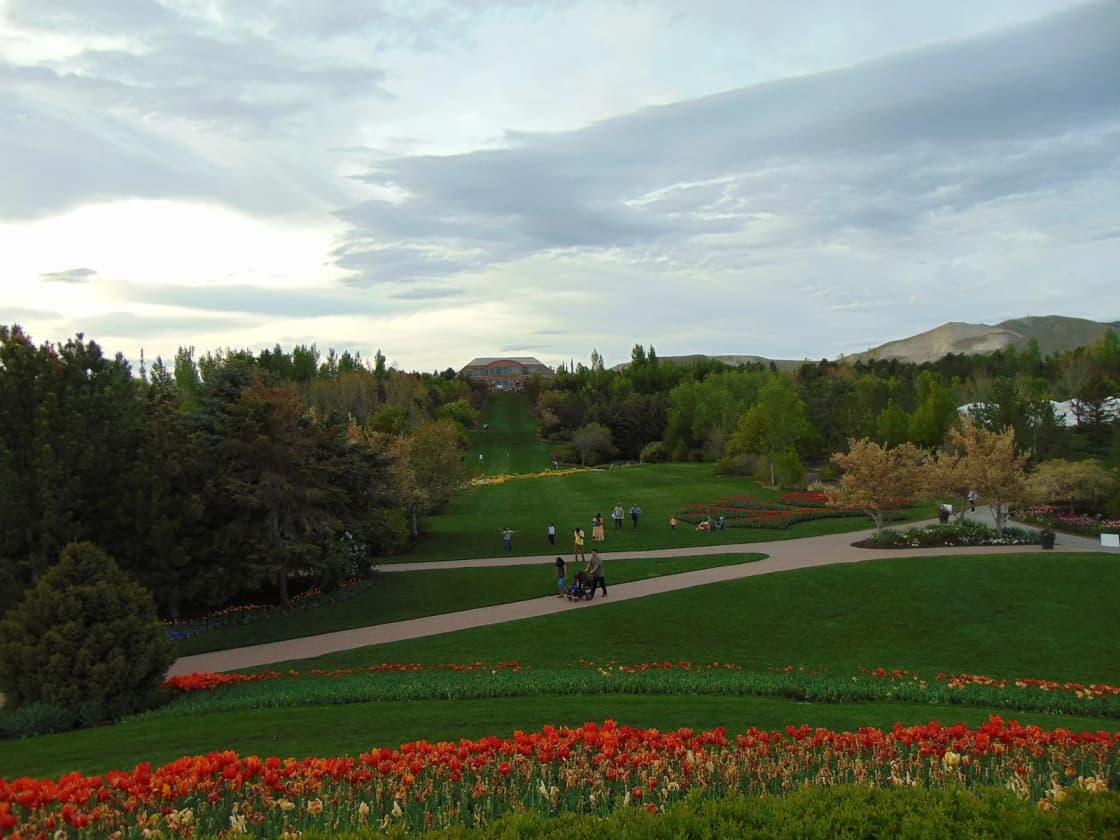 Looking north across the lawns and red tulip beds of Ashton Gardens at Thanksgiving Point in Lehi, Utah, toward the visitors center under a cloudy sky