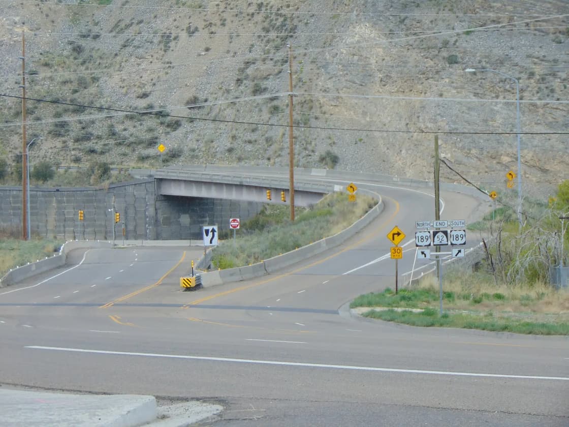 The road junction at the mouth of Provo Canyon where U.S. 189 meets State Route 52, with route signs, a concrete overpass, and a dry brown hillside rising behind, looking east into the canyon