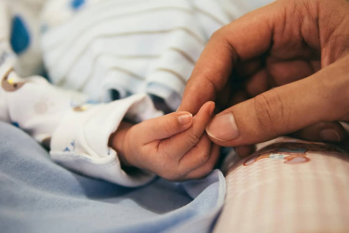 A newborn's small hand wrapped around an adult's index finger against striped hospital-style blankets.