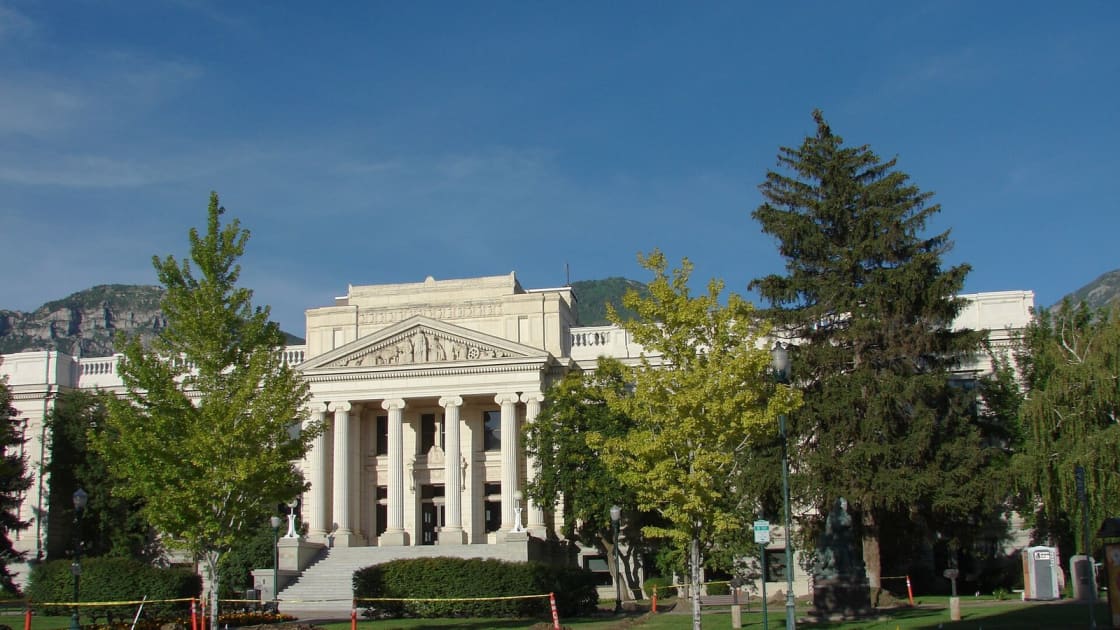 The historic Utah County Courthouse in Provo, a pale stone building with tall columns and a sculpted pediment, framed by trees with mountains behind