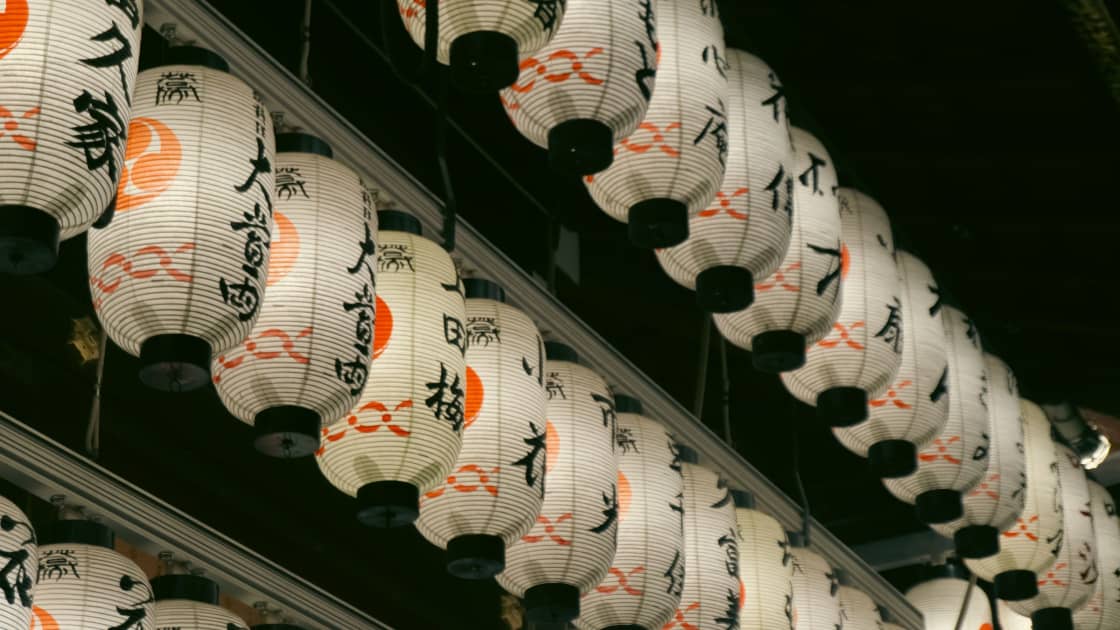 Rows of white paper lanterns hung in tiers under a dark eave, each lit from inside and painted with black calligraphy, an orange disc and an orange knotted cord motif