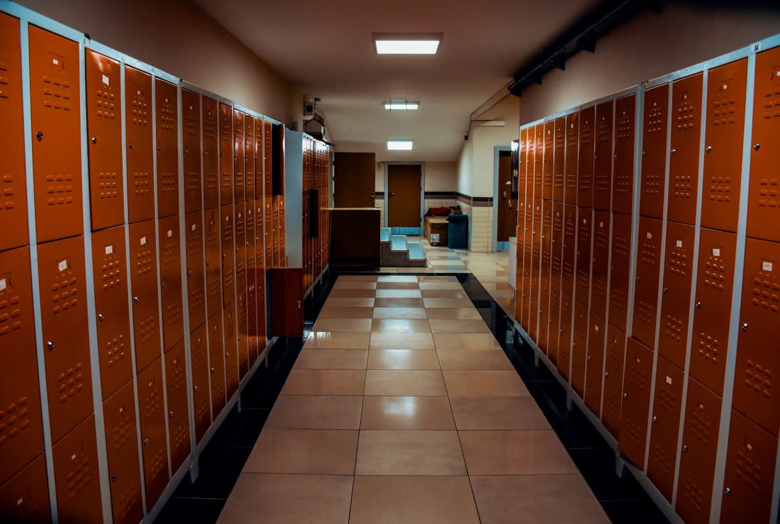 A corridor lined on both sides with tall orange metal lockers above a tiled floor, lit by ceiling panel lights