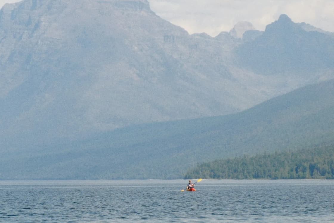 A lone kayaker in a red boat on calm, misty water, with steep gray mountains rising across the lake
