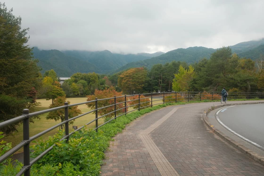 A paved path with a wooden rail fence curves past green trees toward cloud-topped mountains
