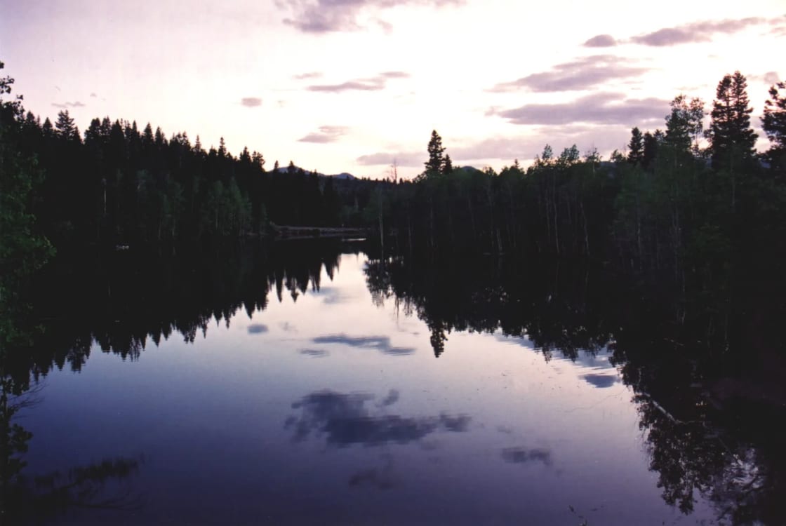Still water at dusk on one of the Payson Lakes, with dark stands of fir and aspen on both shores and a pale lavender sky and clouds reflected in the lake