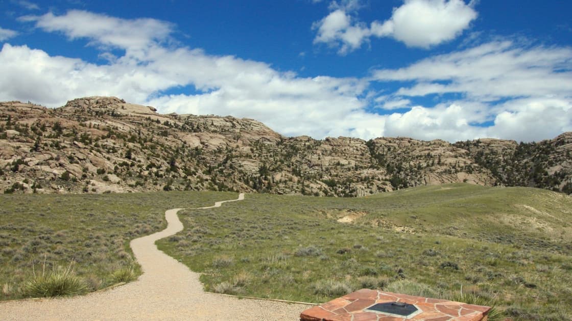 The trail to Martin's Cove in Wyoming winding through sagebrush and grass toward the rounded granite hills of the cove under a blue sky with white clouds