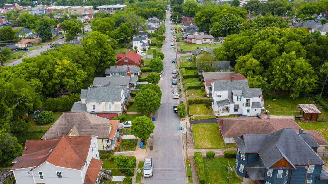 An aerial view along a residential street of older gabled houses with deep mature tree canopy on both sides and cars parked at the kurb