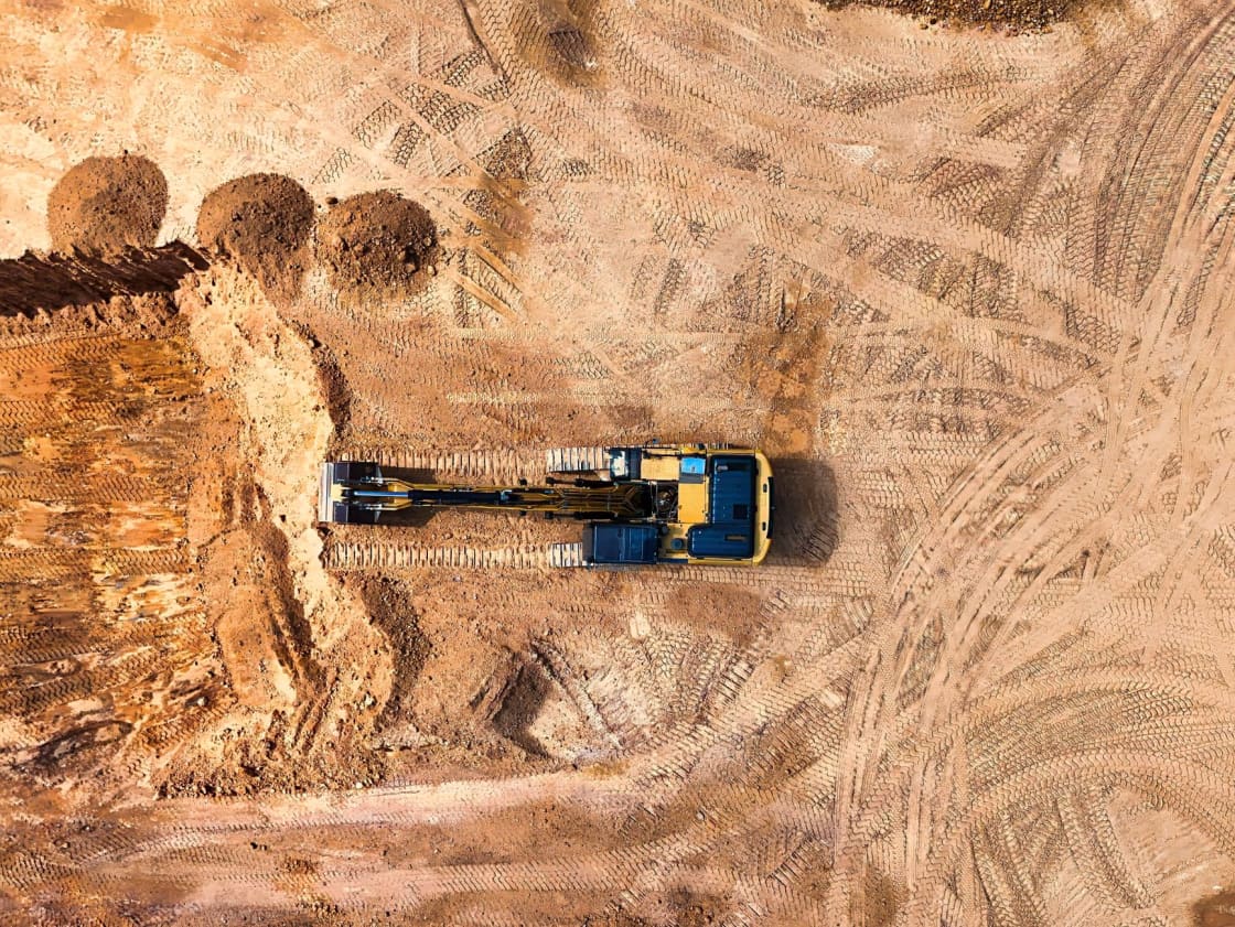 Overhead aerial view of a yellow tracked excavator parked on bare graded earth criss-crossed with tire tracks at a construction site
