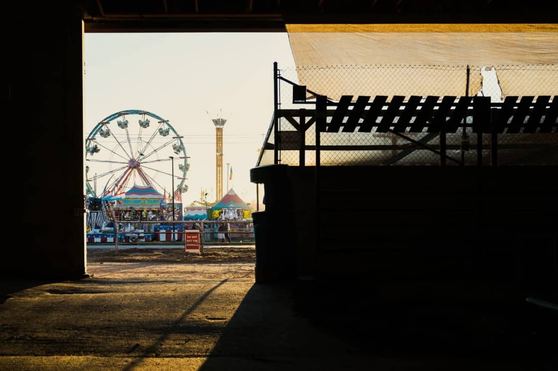 A fairground ferris wheel and swing tower seen through a dark gap between two structures at dusk
