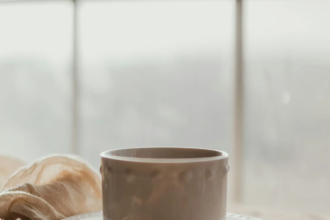 A ceramic cup on a saucer rests beside a rumpled blanket at a bright, fogged window, a quiet moment at home.