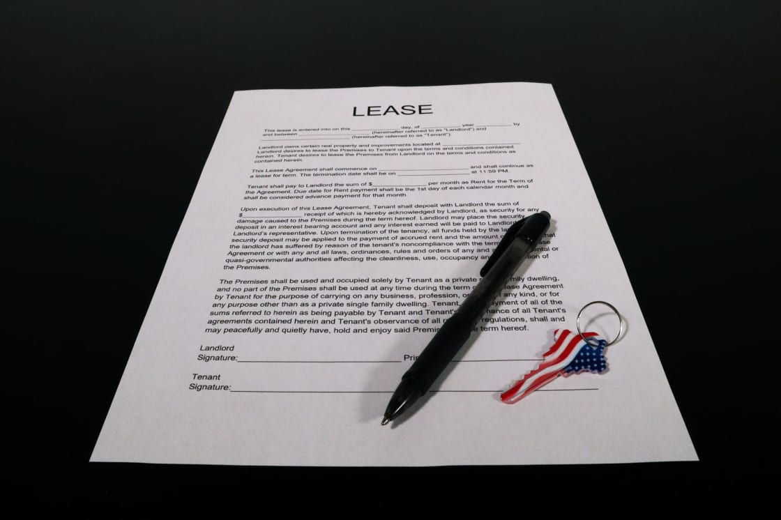 A blank printed lease agreement with landlord and tenant signature lines, a ballpoint pen and a key with a United States flag pattern on a dark surface