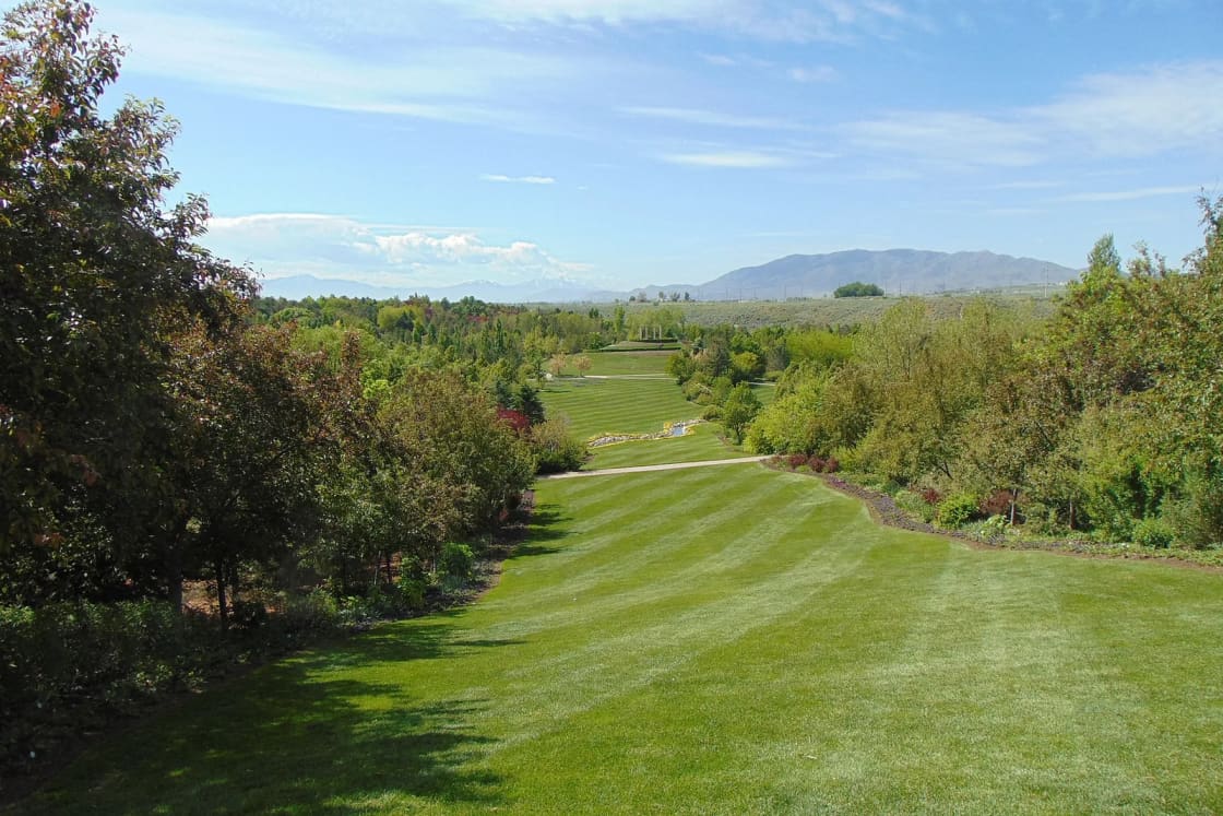 A long striped lawn running between trees and flower beds at Ashton Gardens in Lehi, with mountains in the distance.