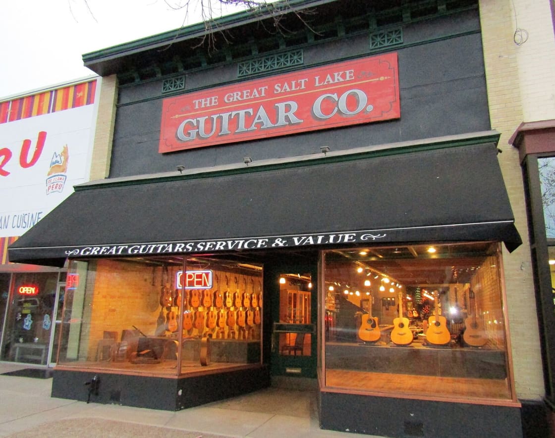 A guitar shop storefront on a downtown Provo street lit at dusk, acoustic guitars in the window behind a glowing neon OPEN sign