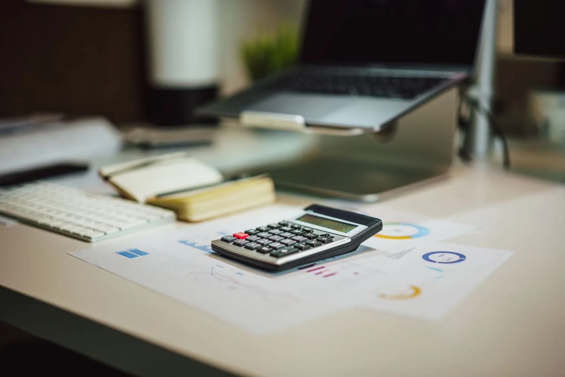 A calculator, notepad and printed financial charts on a desk beside a laptop