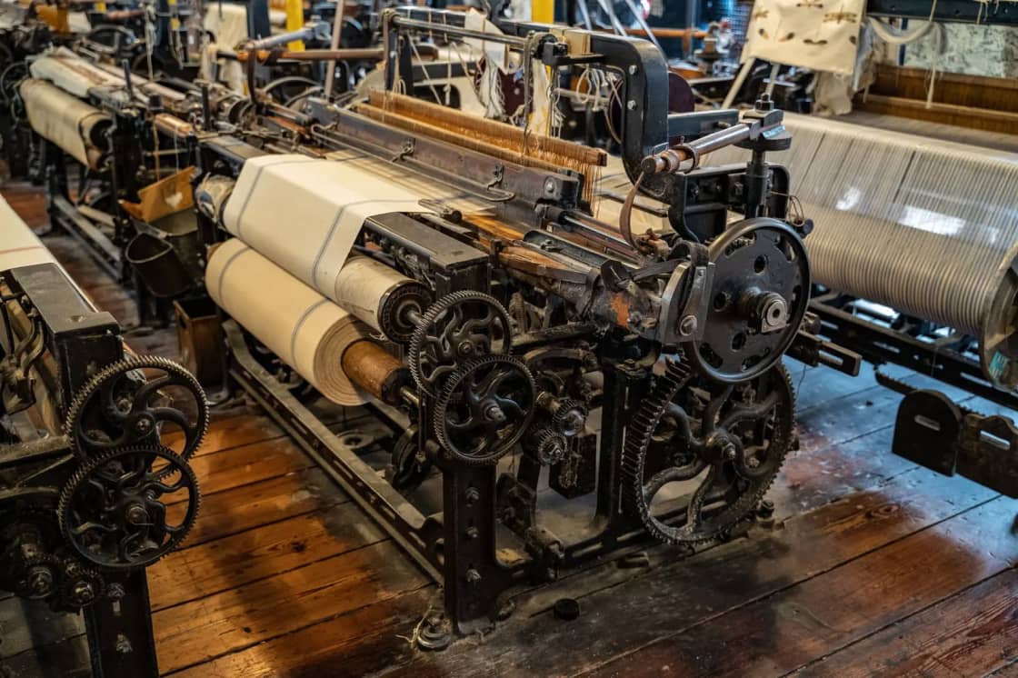 An antique cast-iron power loom with pale woven cloth wound on its beam, standing on a worn plank floor
