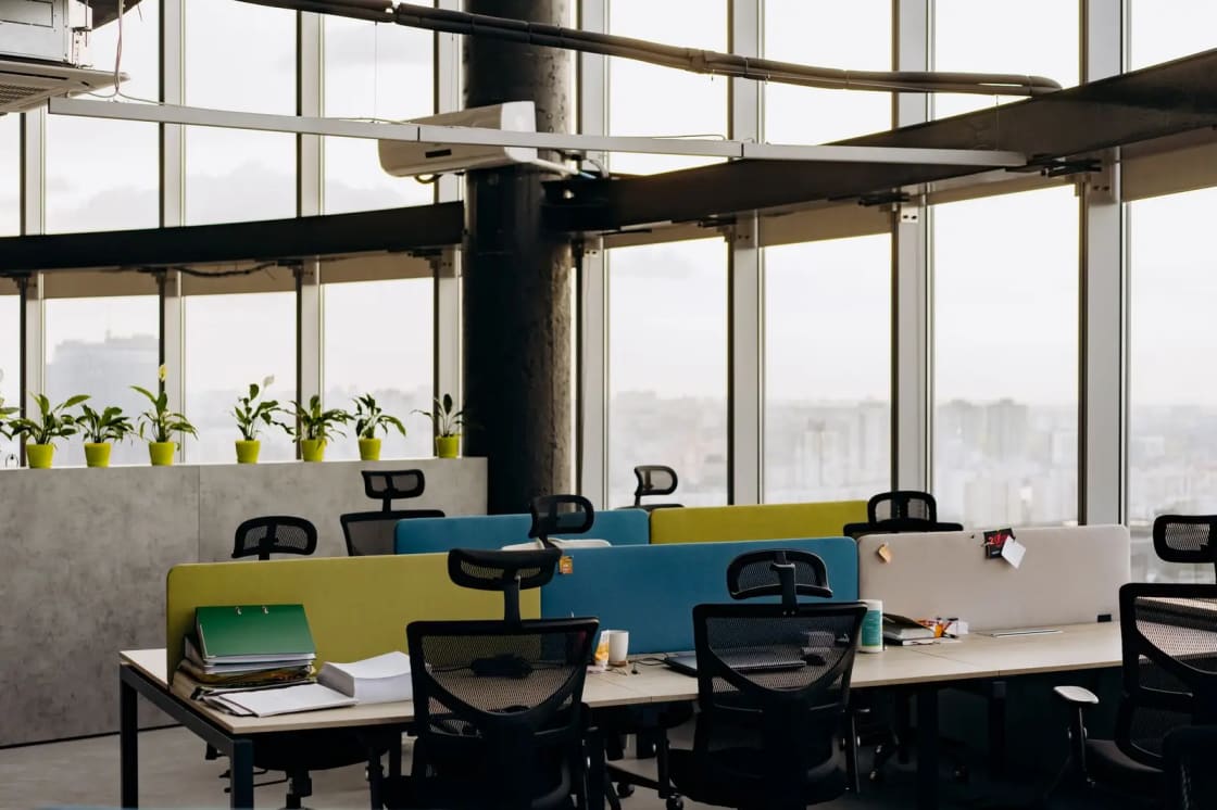 An open-plan office with empty desks and chairs beside a wall of windows