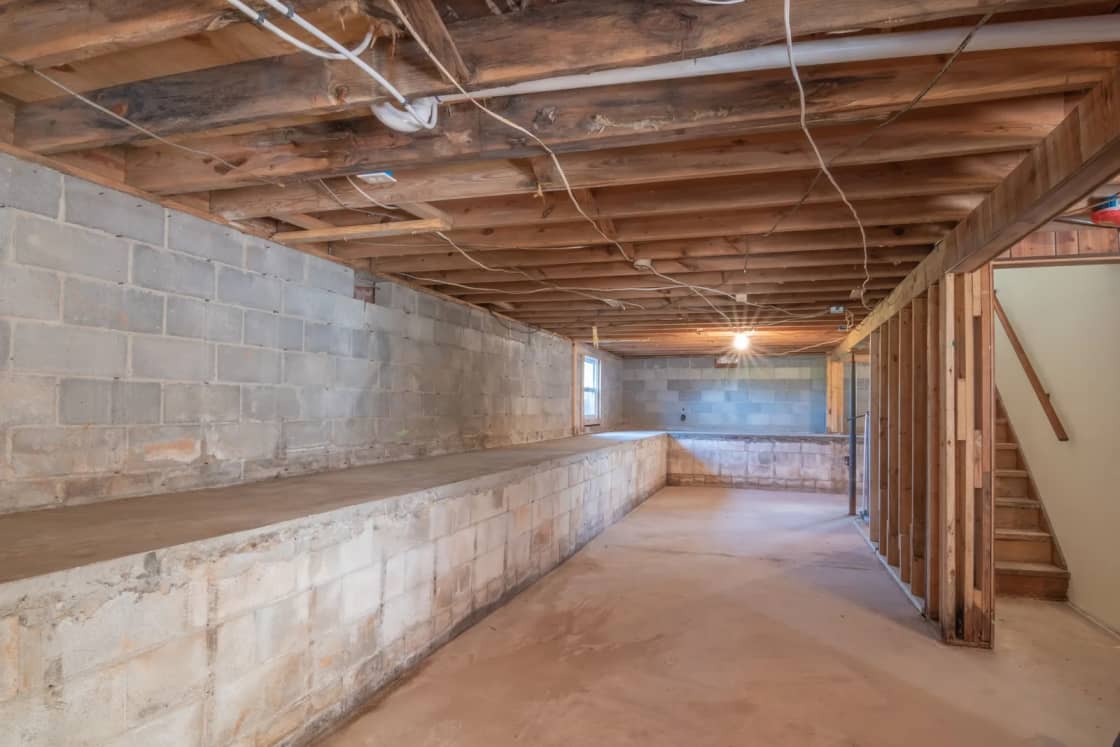 An unfinished basement with concrete block walls, exposed joists overhead and a wooden staircase.