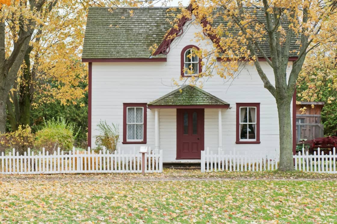 A white two-story farmhouse with red shutters and a red front door behind a white picket fence, under maples in fall color