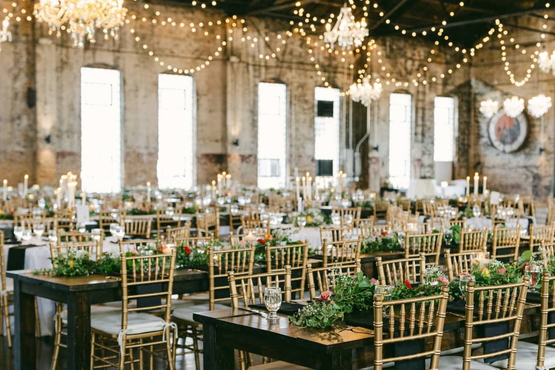 A converted warehouse reception hall set with long tables, gold chairs and greenery under crystal chandeliers and strings of lights.