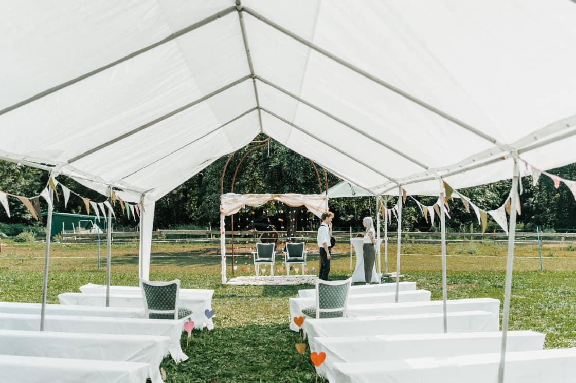 A white frame marquee tent pitched on a lawn and strung with bunting, with rows of white-draped seating and a decorated arch at the far end