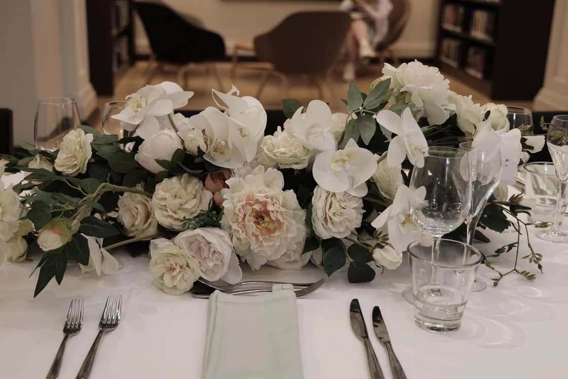 A reception table with a long centerpiece of white roses and orchids beside glasses and silverware.