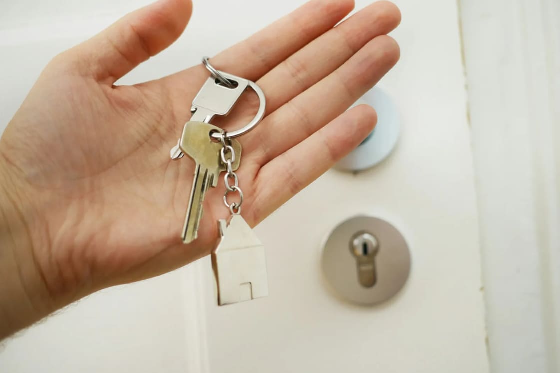 An open palm holding two house keys on a ring with a small tag, held out beside the round deadbolt and knob of a white front door