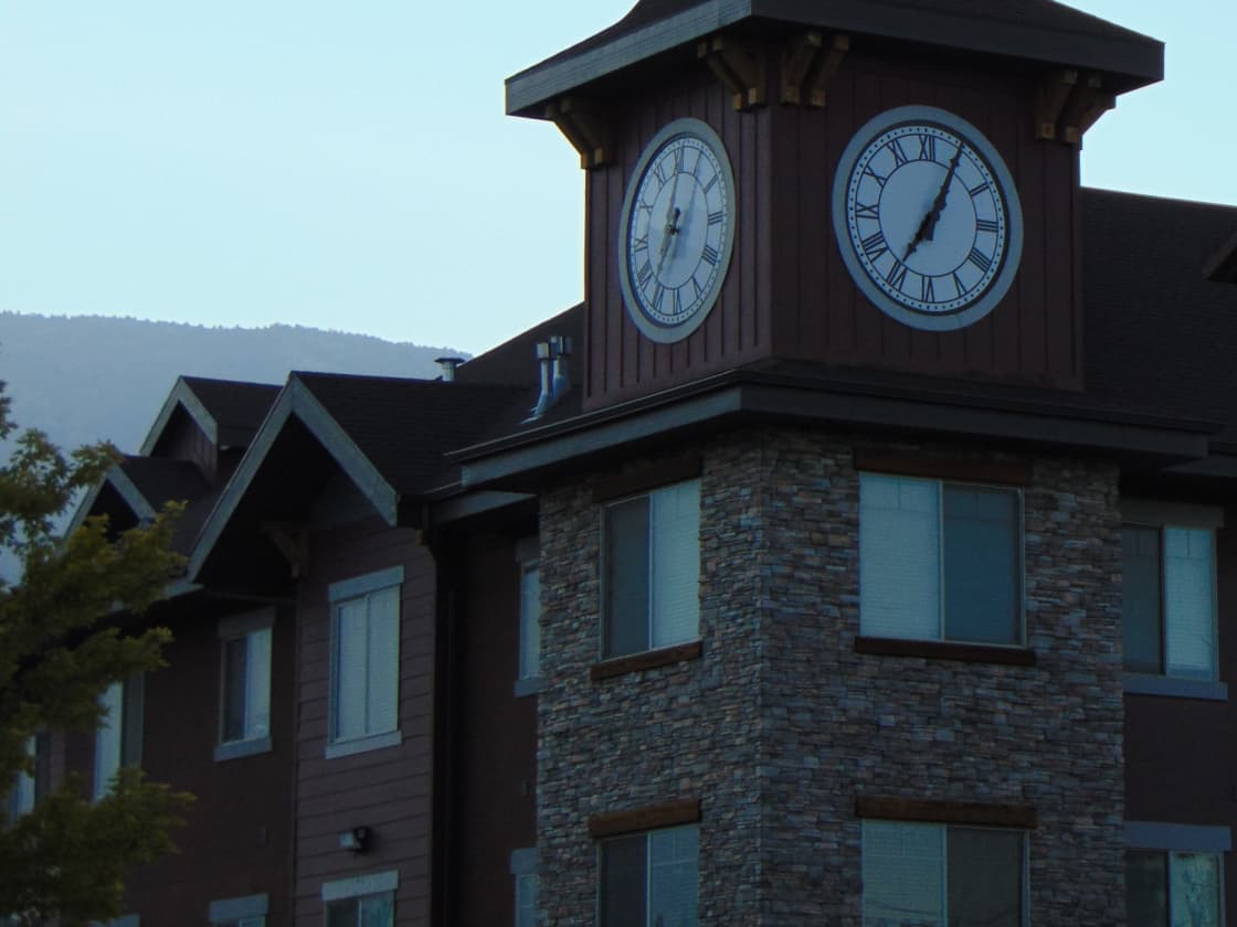 A multi-story apartment building with stone-clad corner, gabled roofs and a clock tower, with a wooded hillside behind