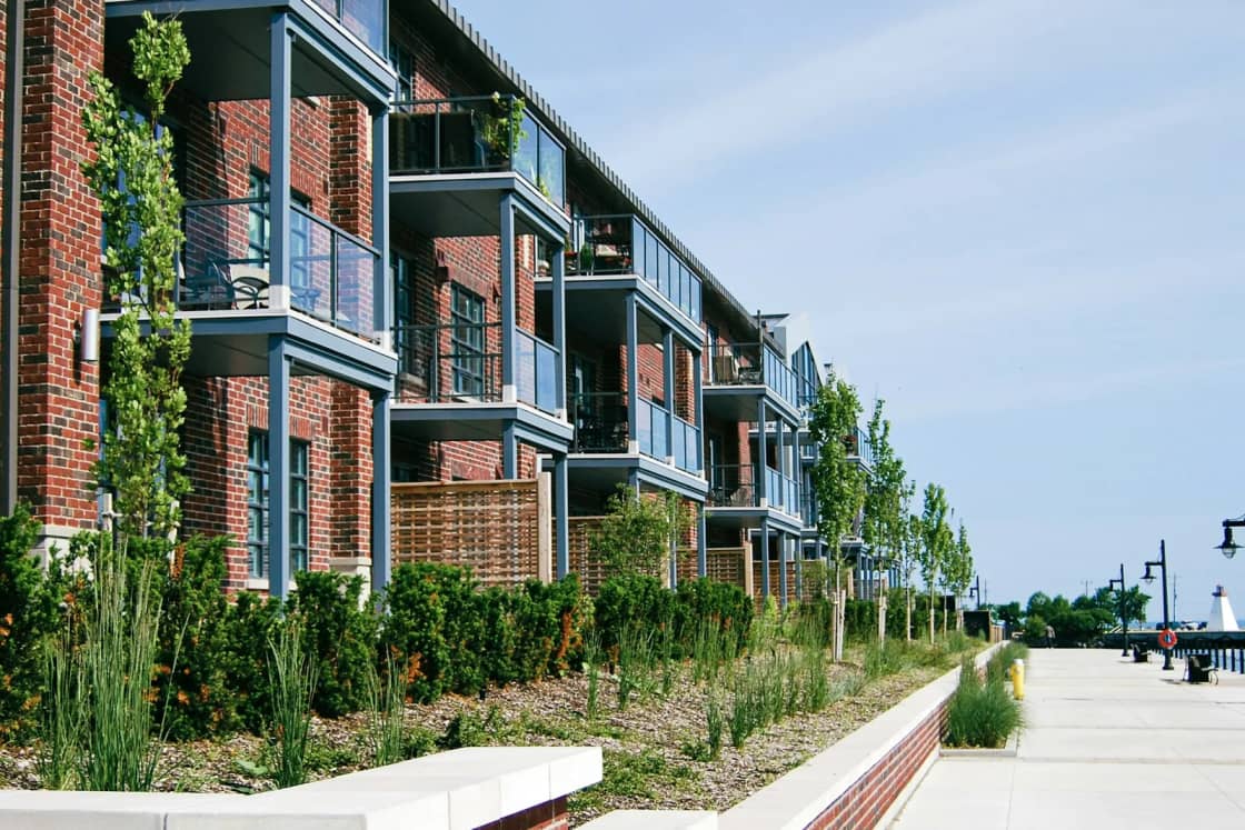 A long red-brick apartment building with stacked balconies and climbing vines along a wide sidewalk on a sunny day