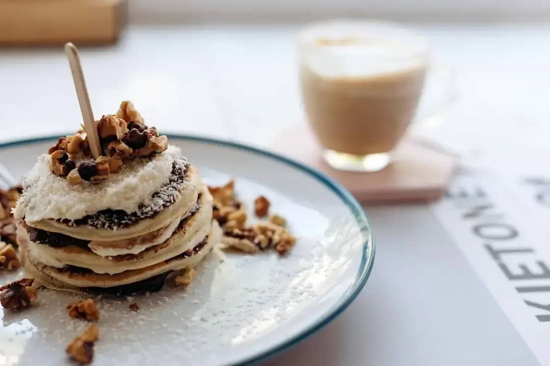 A stack of pancakes topped with cream and nuts beside a latte