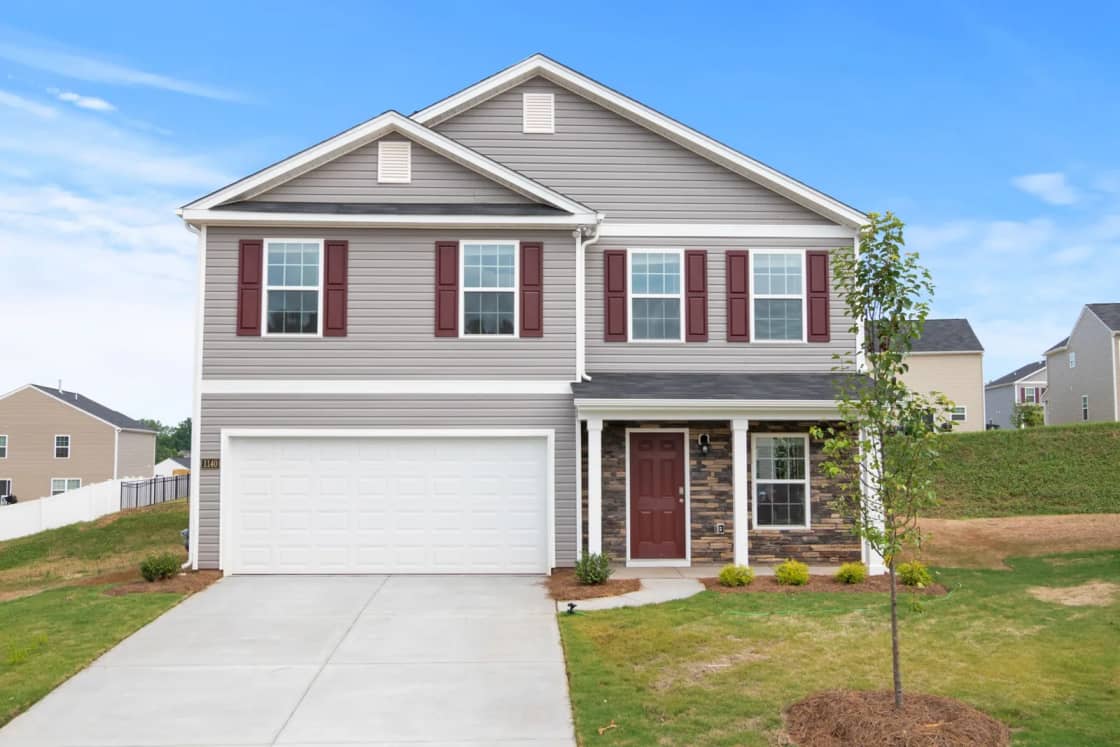 A new two-story house with maroon shutters, a white garage door and a young tree in the front lawn.