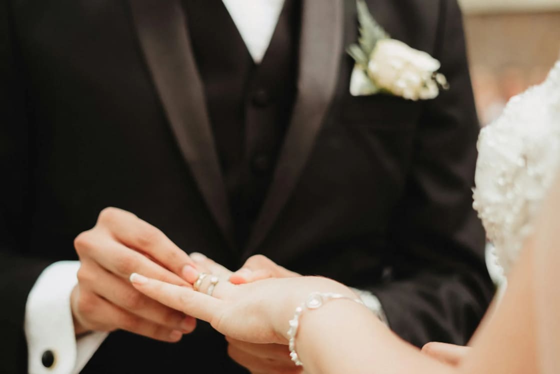 A groom in a black tuxedo with a white rose boutonniere slides a ring onto his bride's finger under strings of warm lights.