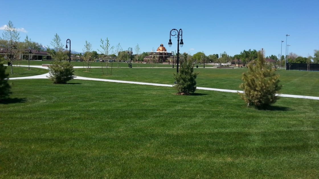A wide green lawn in Riverton City Park with young pines, curving paths and black lamp posts, and a domed brick building in the distance under a clear blue sky