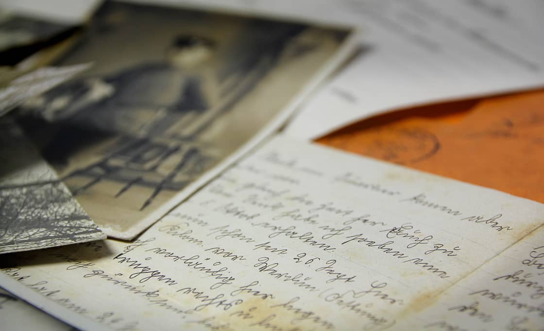 A close-up of old family papers: two foxed handwritten letters in looping cursive, a small black-and-white photograph of a figure standing outdoors resting on top of them, and the corner of an orange envelope carrying a faded postmark