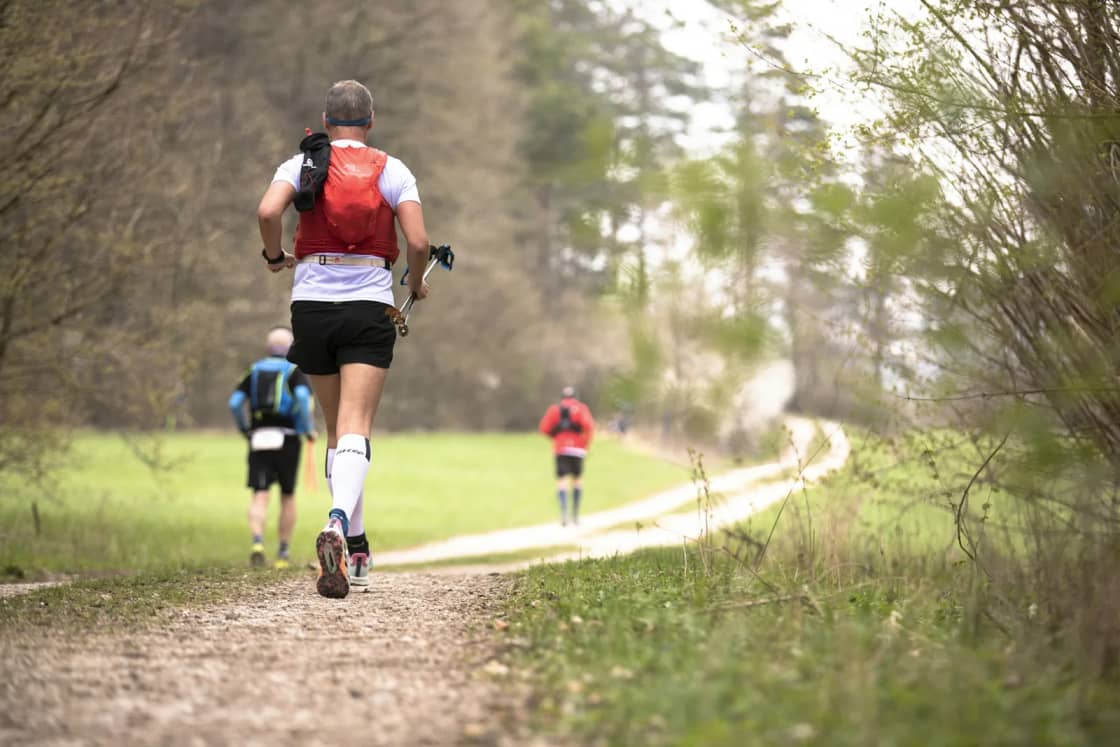 A few runners seen from behind on a dirt path through spring woods, the nearest wearing an orange hydration vest
